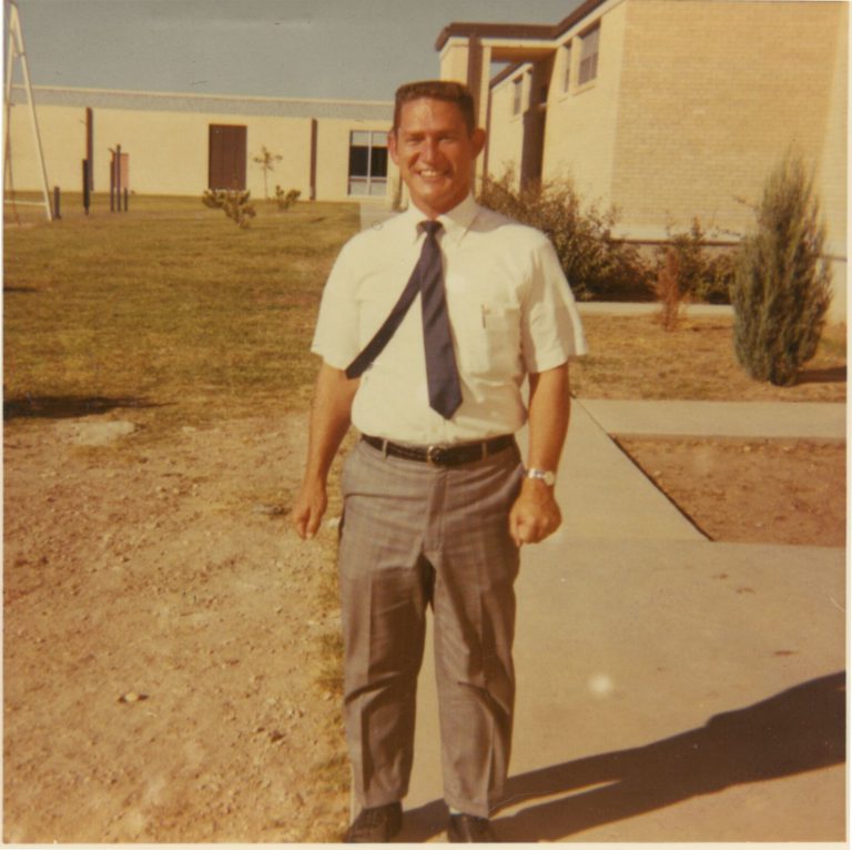 Coach Jimmie Keeling of the 1968 Estacado Matadors Football team smiling in a short-sleeved shirt and tie outside a building