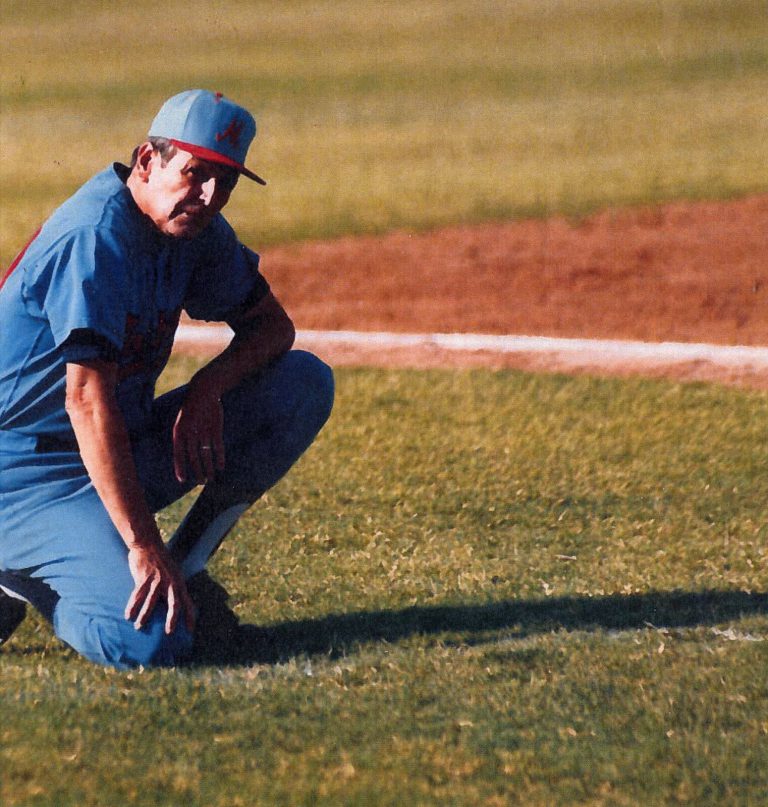 Coach kneeling on the grass in a light blue uniform and M cap, from Bobby Moegle's collection