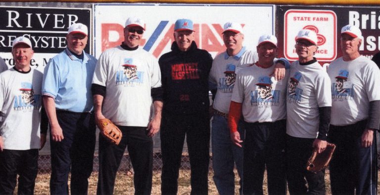 Bobby Moegle in a Monterey Baseball sweatshirt with alumni in matching T-shirts in front of an outfield wall