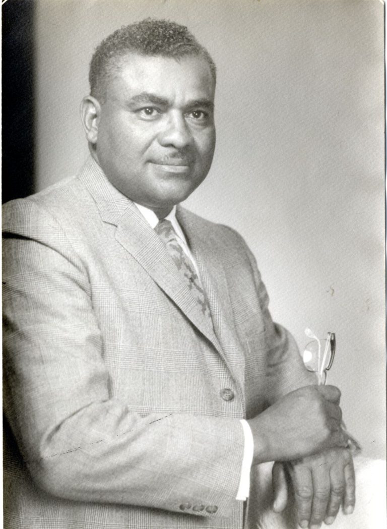 Damon Hill, Sr. in a light plaid suit jacket and patterned tie, holding eyeglasses, in a black-and-white studio portrait