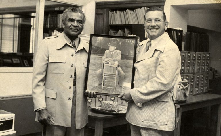 Two men in light jackets holding a framed photograph of a man in a lawn chair in a library, Damon Hill, Sr. collection