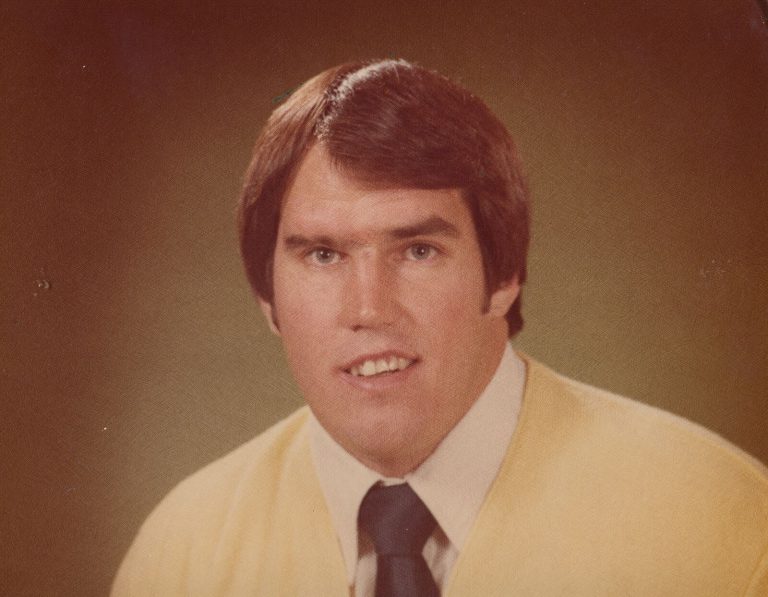 Dan Irons in a yellow cardigan and dark tie, color studio portrait