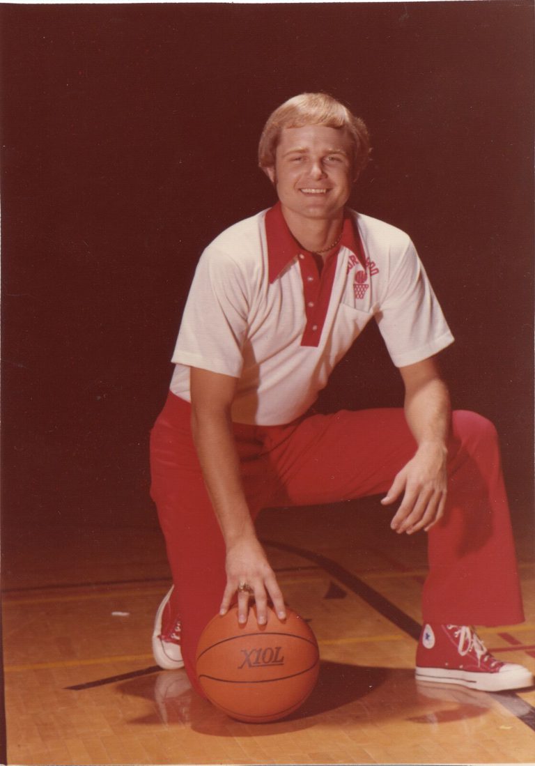 David Thetford in a red and white polo and red slacks, kneeling with a basketball on a court