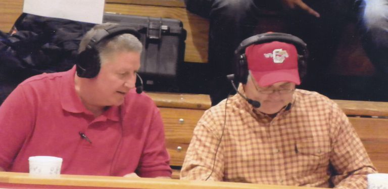 David Thetford's radio career: two men in headsets at a broadcast table in front of gym bleachers