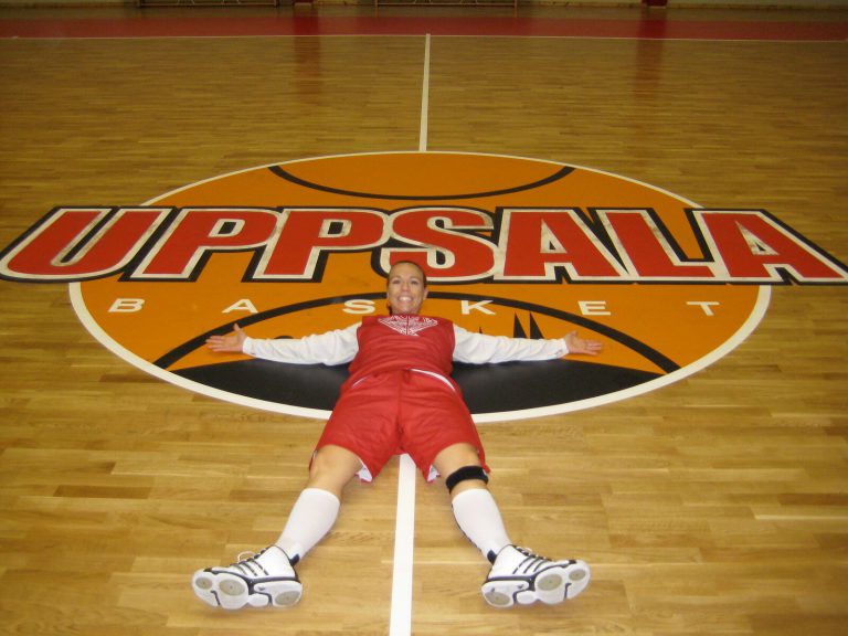 Erika Valek smiling with arms spread, lying on the Uppsala Basket logo at center court