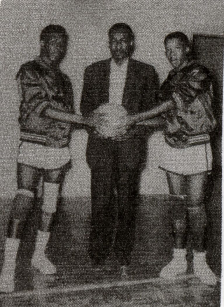 Man in a dark jacket holding a basketball between two players in satin warm-up jackets, from Ernest Mallory's scrapbook