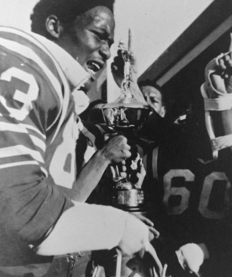 Fred White, number 83, holding a trophy and shouting with joy, with a teammate's number 60 jersey behind
