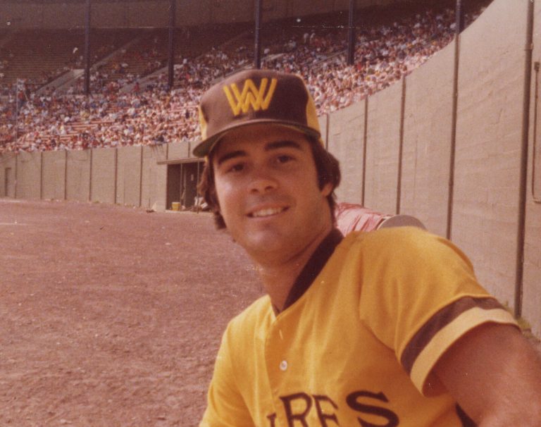 Gary Ashby smiling in a gold baseball uniform and W cap in front of a ballpark grandstand, color snapshot