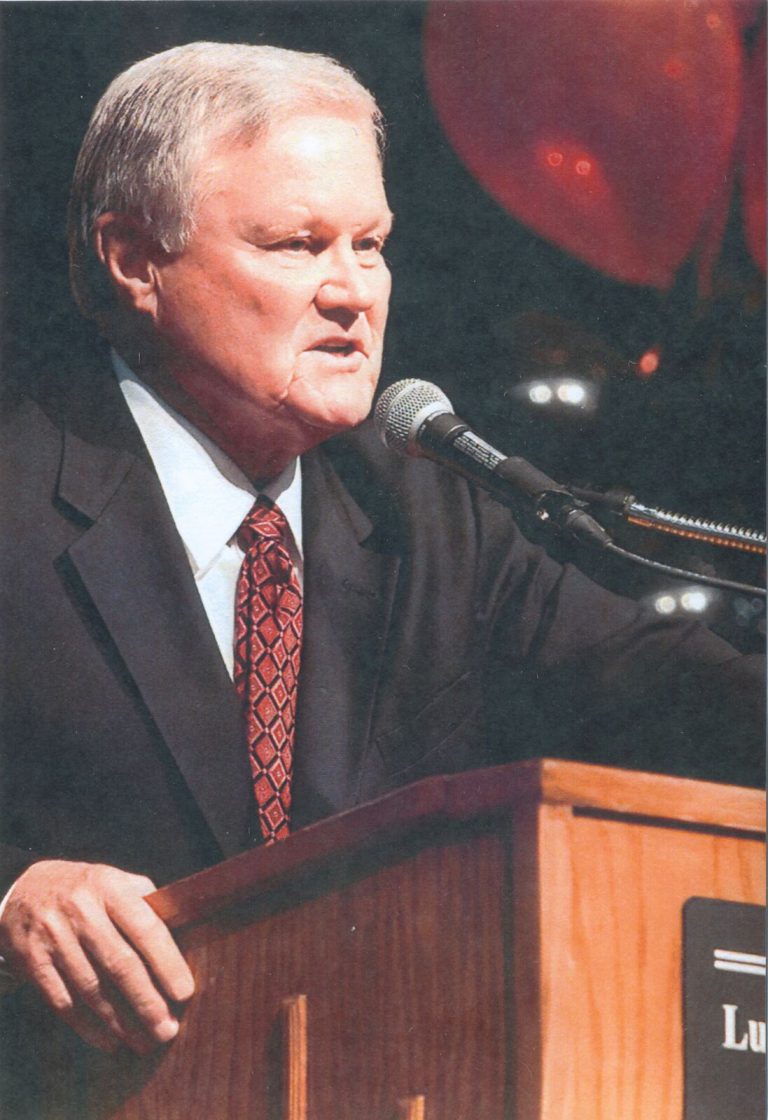 Gerald Myers speaking at a wooden podium with a microphone, red balloons in the background