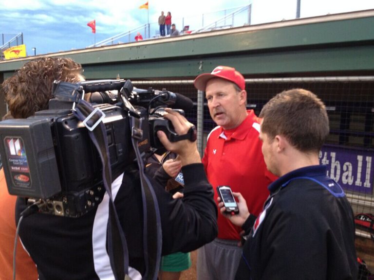J. J. Johnson in a red polo being interviewed by a TV crew at a softball field