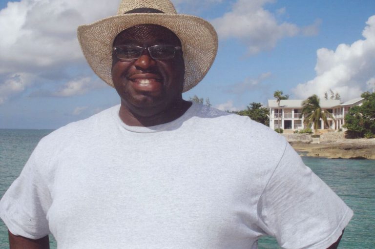 James Collins smiling in a straw hat and glasses by the water, with buildings across the bay behind