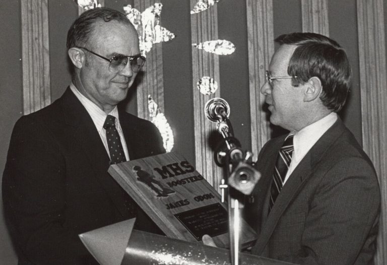 James Odom, in tinted glasses and a dark suit, receiving a wooden MHS Boosters plaque from a man at a microphone