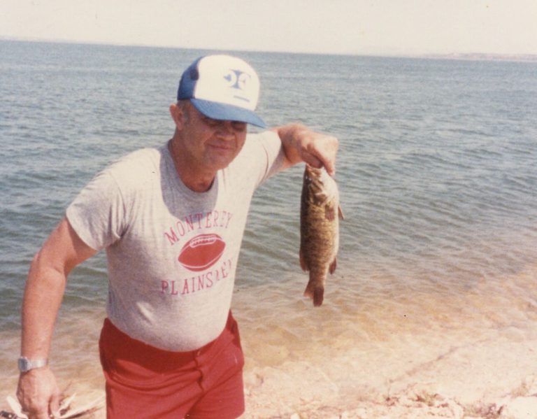 James Odom holding up a fish beside a lake, in a Monterey Plainsmen shirt and a cap