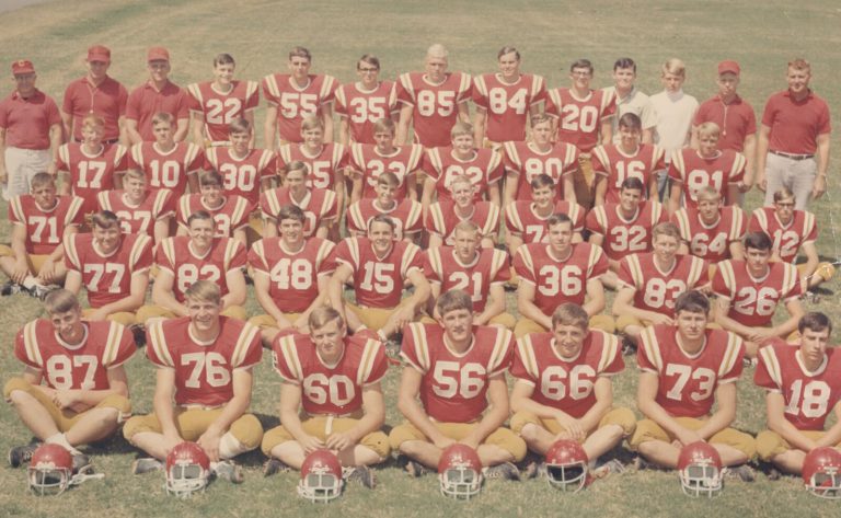 Football team in red jerseys posed in rows with coaches and helmets in front, from Jerry Austin's collection