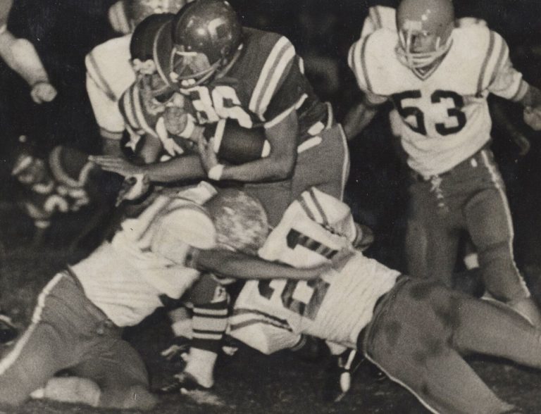 Jerry Austin carrying the football through a crowd of tacklers in a black-and-white night game photo