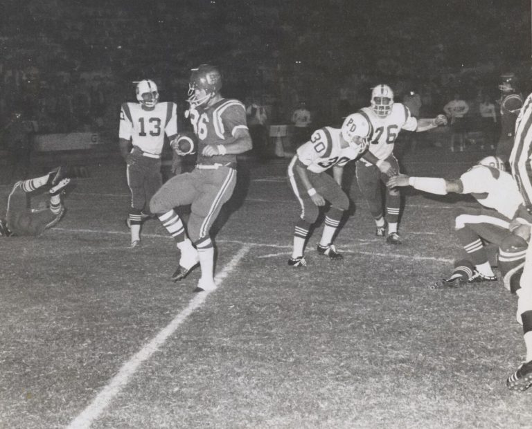 Jerry Austin, No. 36, running upfield with the football past defenders in a black-and-white night game photo