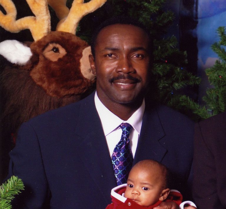 Jerry Gray in a navy suit and patterned tie holding a baby in red beside a plush reindeer and a Christmas tree
