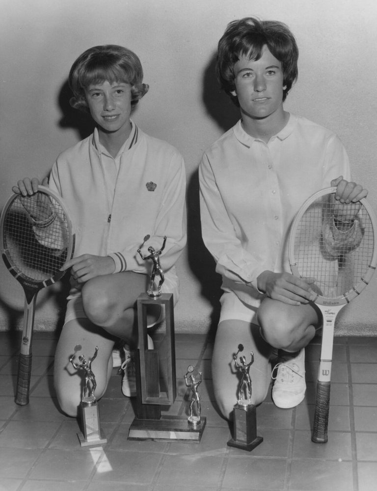 Jill Philbrick Gentry kneeling with a teammate beside tennis rackets and trophies