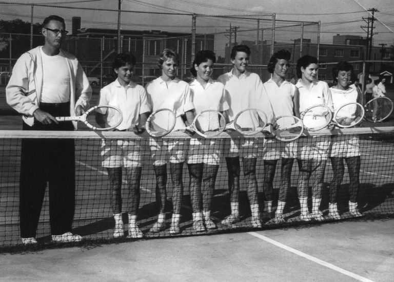 Jill Philbrick Gentry's tennis team lined up at the net with rackets beside a man in a white jacket