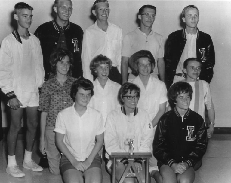 Tennis team and coach posed indoors with a trophy, several in L letter jackets, from the Jill Philbrick Gentry collection