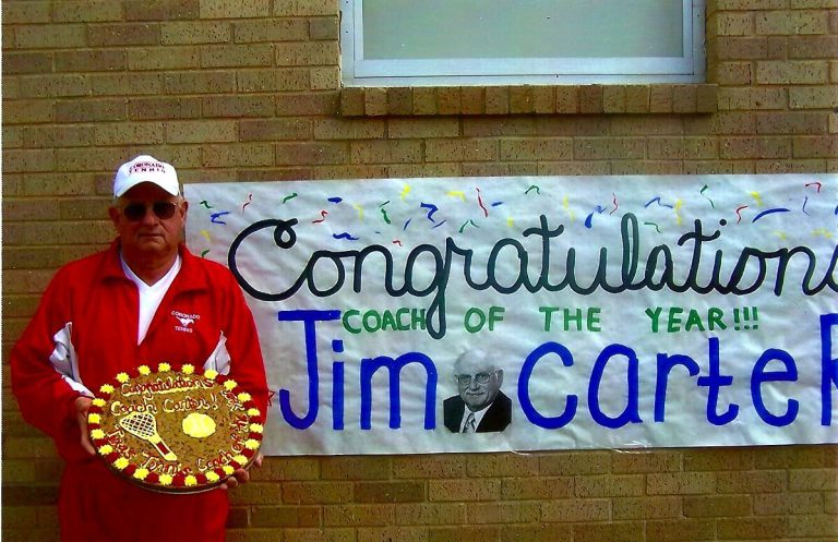 Jim Carter in a red jacket and white cap holding a decorated Coach of the Year cookie beside a congratulations banner