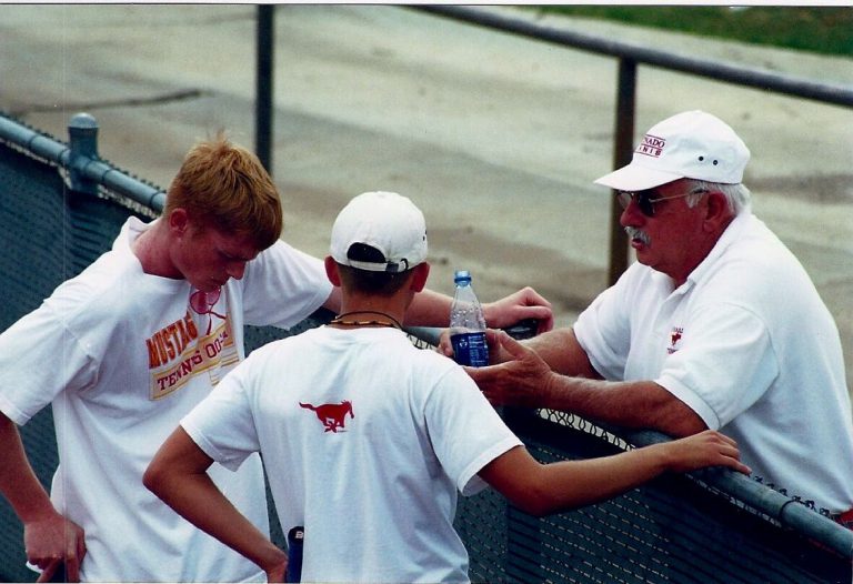 Man in a white Coronado Tennis cap and polo talking with two players at a courtside fence, from Jim Carter's collection