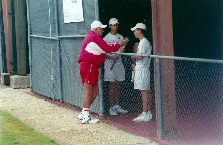 Man in a red jacket and shorts talking with two players in white caps at a court fence, from Jim Carter's collection