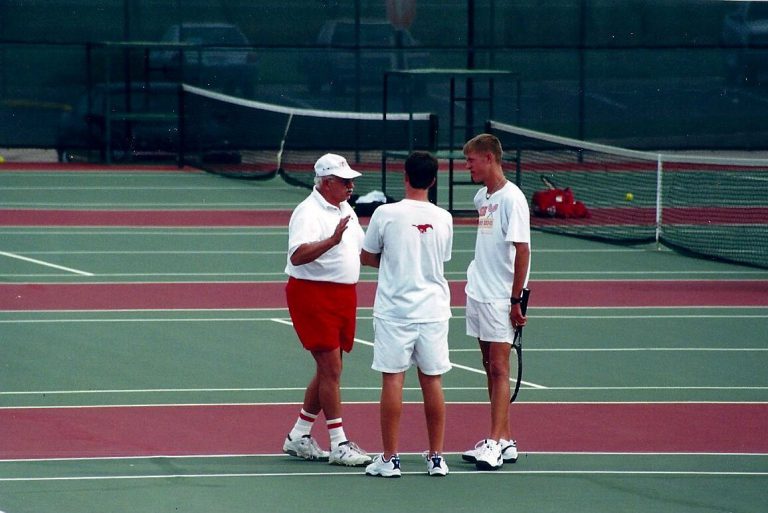 Man in a white cap and red shorts talking with two players in white on a tennis court, from Jim Carter's collection