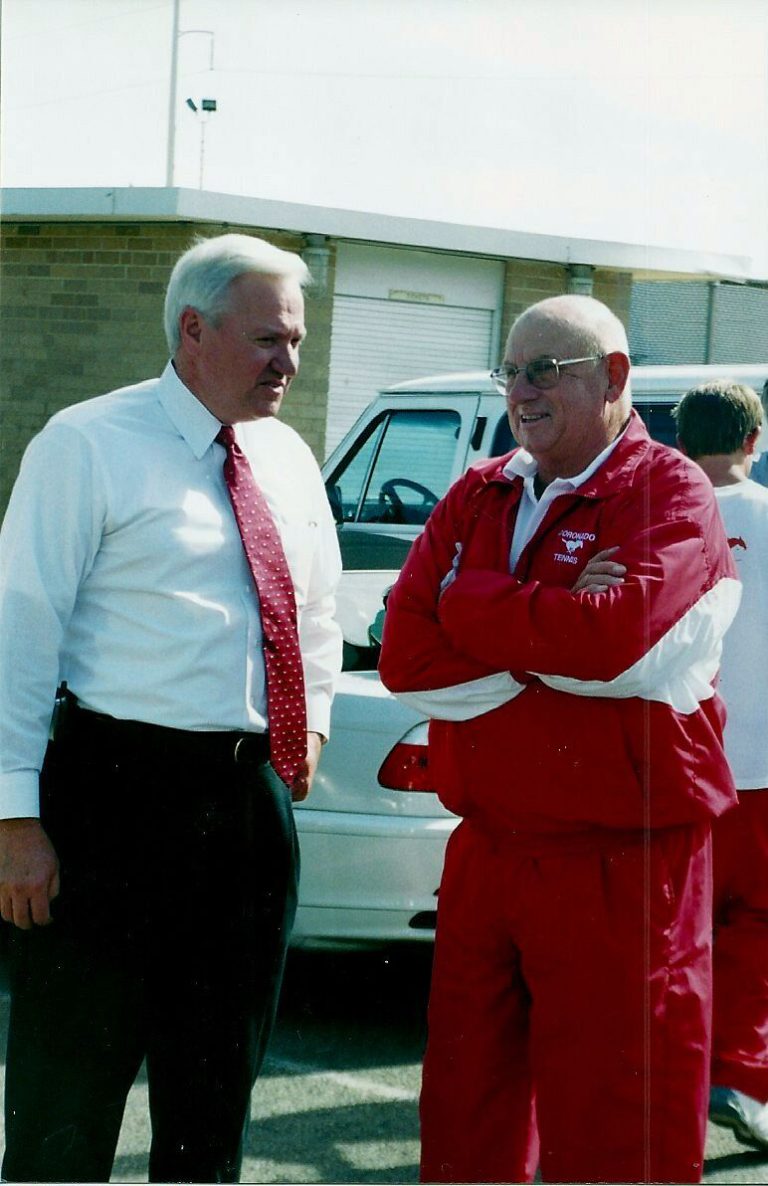 Man in a red Coronado Tennis jacket with arms crossed beside a man in a white shirt and red tie, in Jim Carter's collection