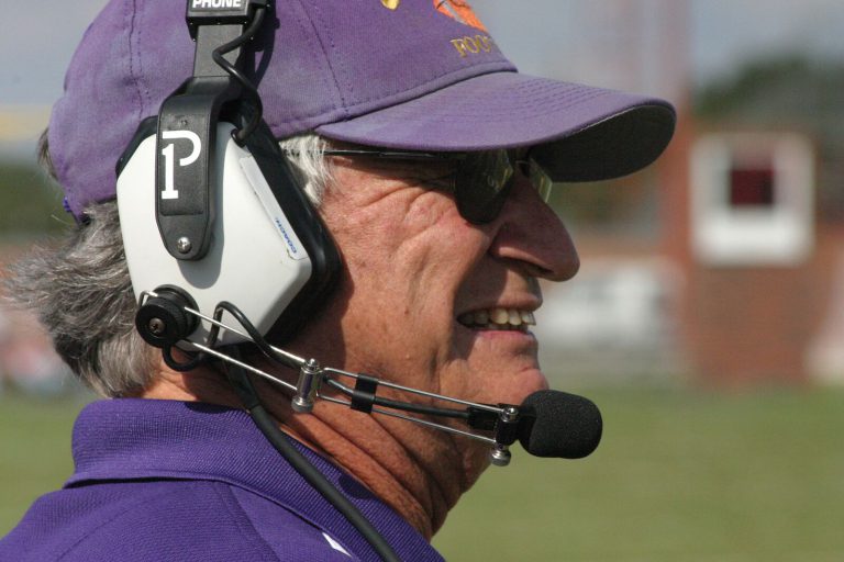 Jimmie Keeling in profile, smiling on the sideline in a purple cap and coaching headset