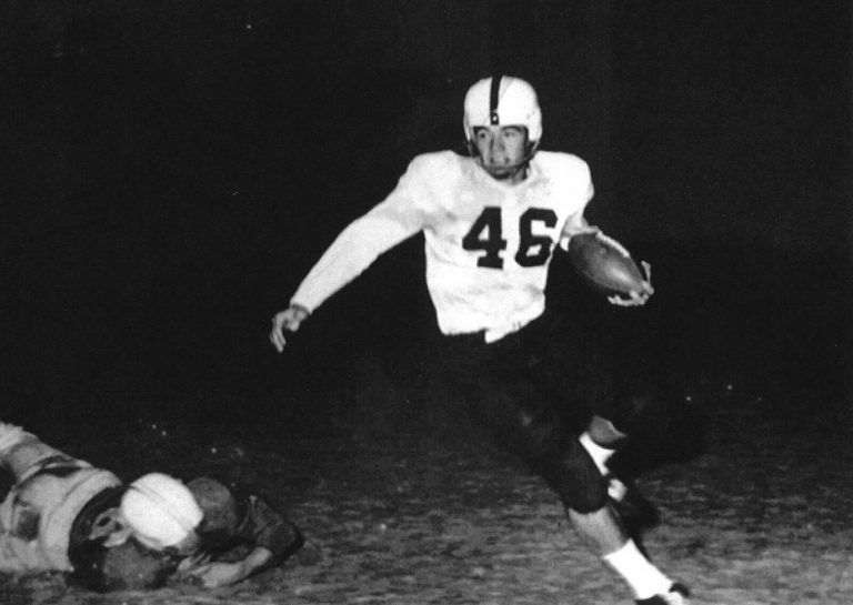 Jimmy Welch, No. 46, carrying the football in a night game as a tackled player lies on the ground