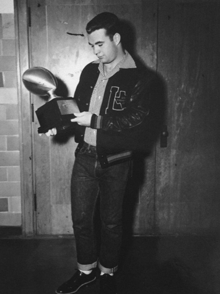 Jimmy Welch standing in a letter jacket and cuffed jeans, looking at a football trophy