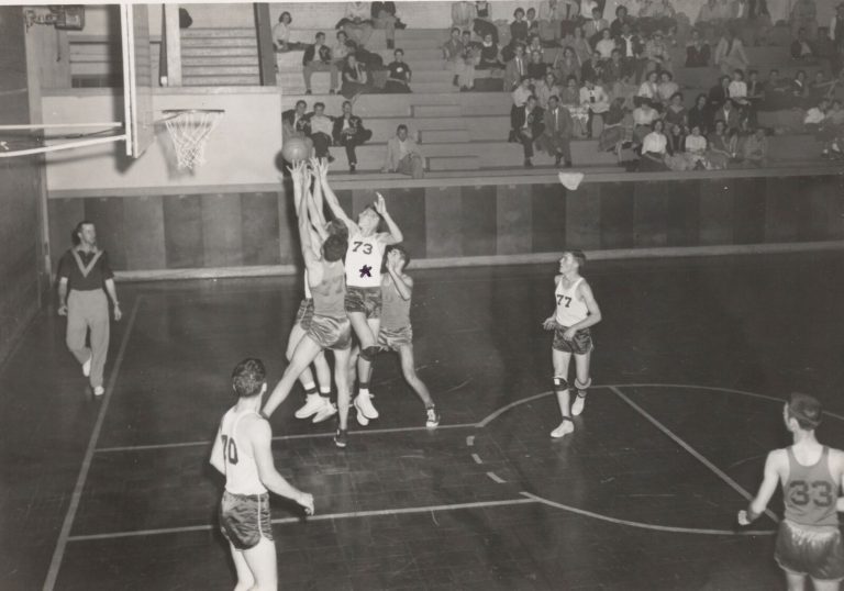 Joe Michalka, number 73, leaping for the ball near the basket among defenders in a gym with spectators