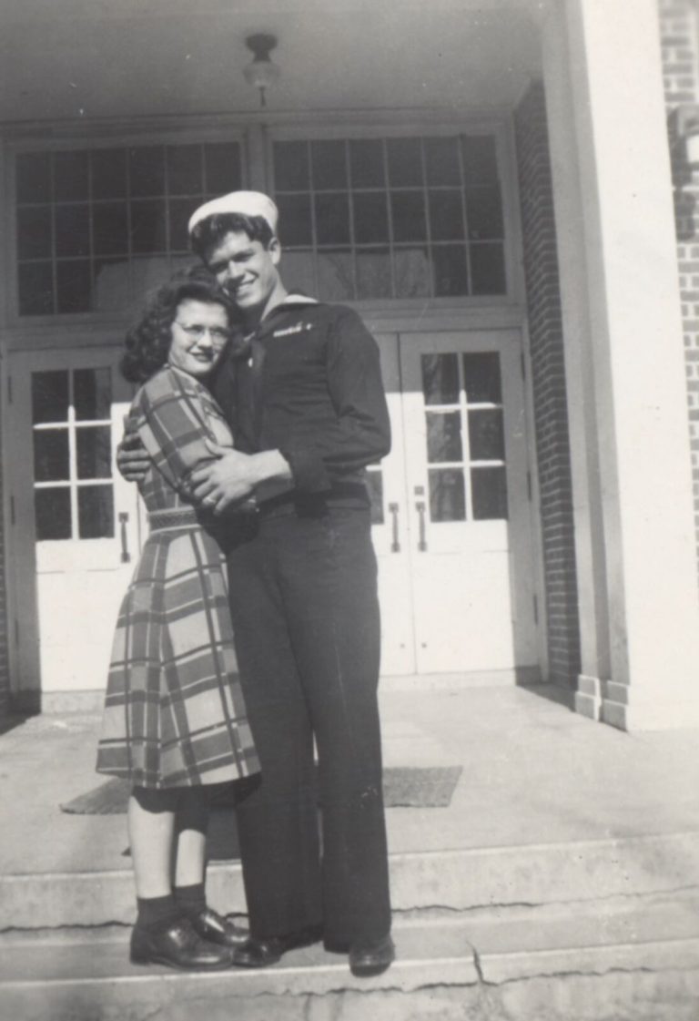 Sailor in uniform embracing a young woman on the steps of a brick building, from the John Cardinal family photos