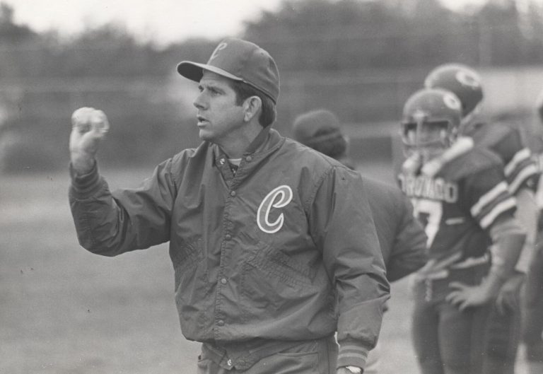John Dudley in a Coronado jacket and cap, gesturing with a raised hand as helmeted players stand behind