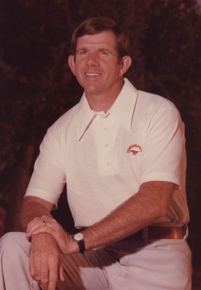 John Dudley smiling in a white polo shirt with a Coronado Mustang logo, kneeling for a color portrait