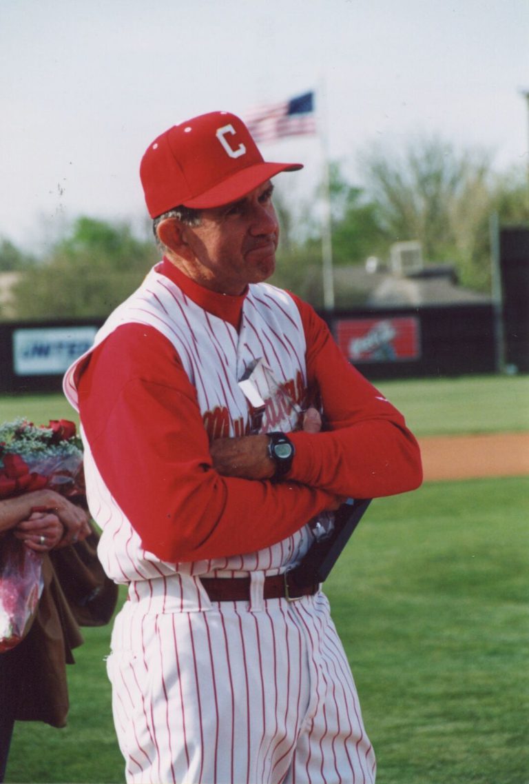 John Dudley in a red cap and red-and-white pinstriped Mustangs uniform, standing on a field with arms crossed