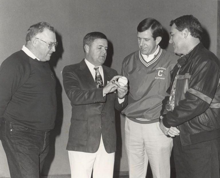 John Dudley, in a Coronado Mustangs pullover, with three other men looking at a baseball in a black-and-white photo
