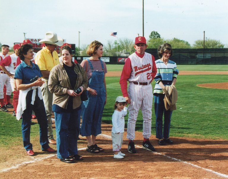 John Dudley in a number 22 Mustangs uniform with a toddler and several adults on the field at a ceremony