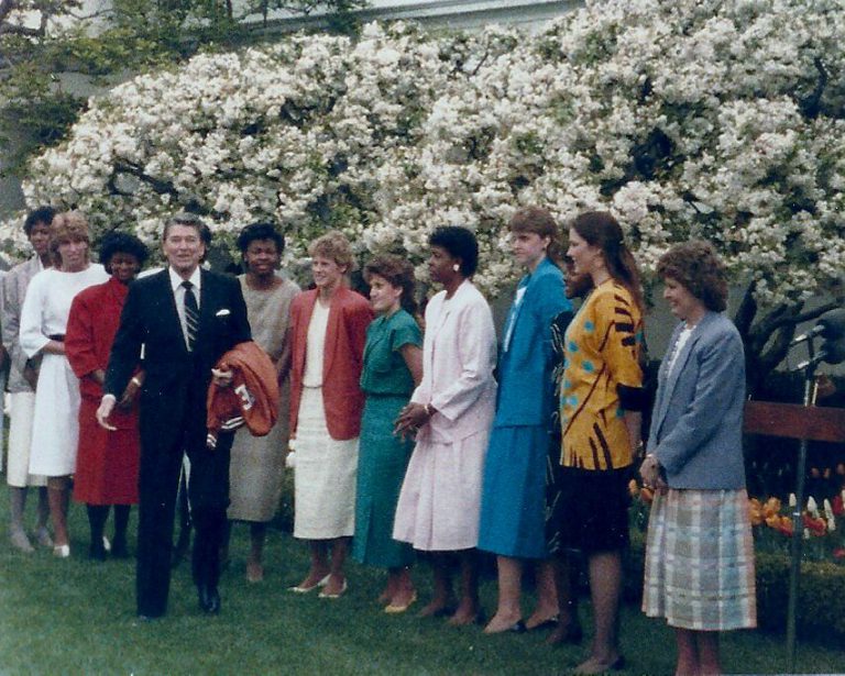Kamie Ethridge's team lined up on the lawn before a flowering tree at the White House beside a man in a dark suit