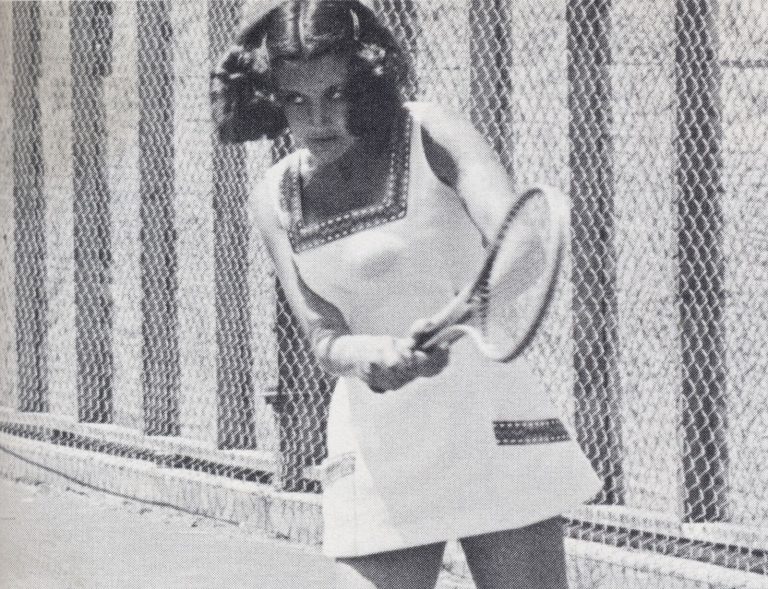 Kathy Kuhne Vick gripping a racket in a white tennis dress beside a chain-link fence, black and white