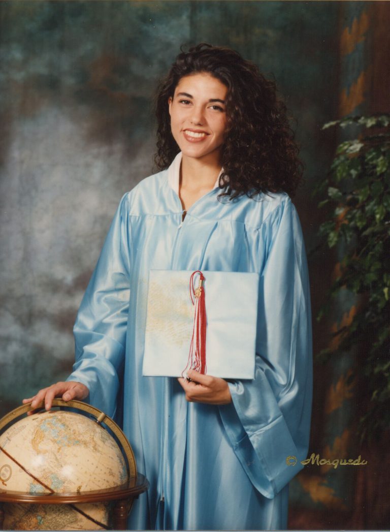 Leigh Daniel Cordes in a light blue graduation gown holding a mortarboard with a red tassel beside a globe