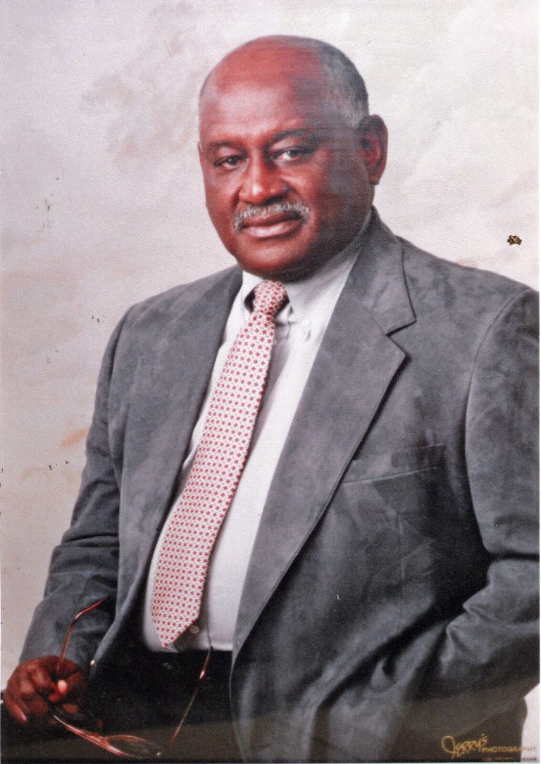 Louis Kelley in a gray suit and patterned tie holding eyeglasses in a studio portrait