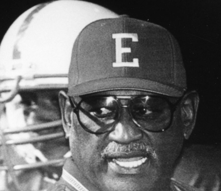 Louis Kelley smiling in a block E cap and dark glasses beside a football helmet in a black-and-white close-up