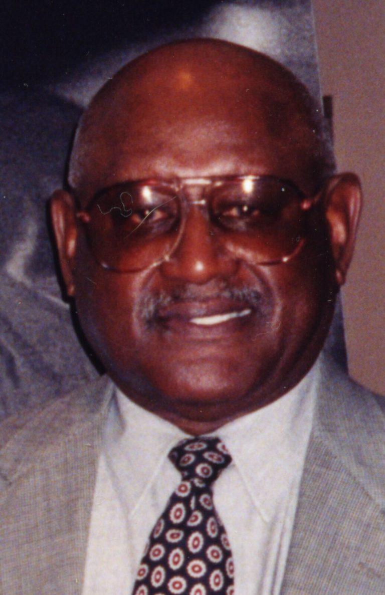 Louis Kelley smiling in tinted glasses, a tan sport coat and a patterned tie