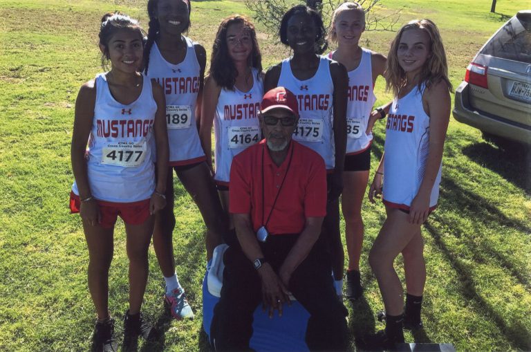 Melvin Houston seated in a red polo with the Coronado Mustangs girls cross-country team standing behind