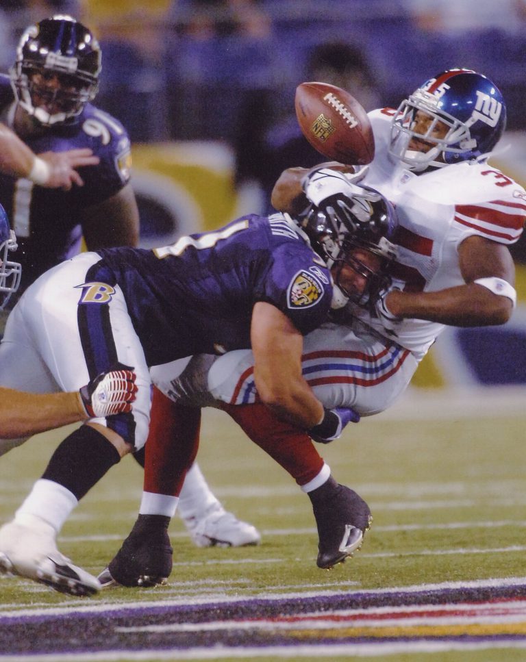 Mike Smith in a purple Ravens uniform hitting a New York Giants player as the ball pops loose