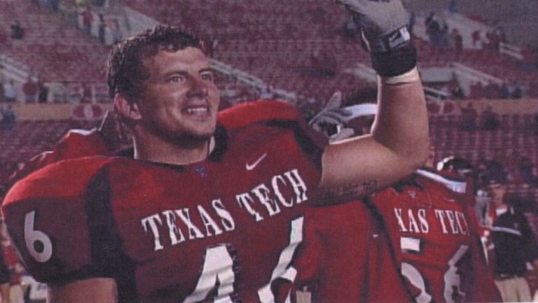 Mike Smith smiling with a gloved hand raised in a red Texas Tech jersey inside the stadium