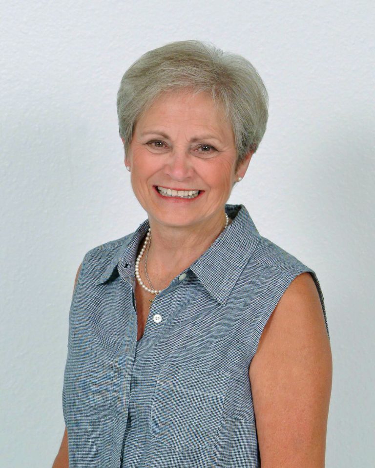 Patty Jones smiling in a sleeveless blue shirt and pearl necklace, portrait against a light wall