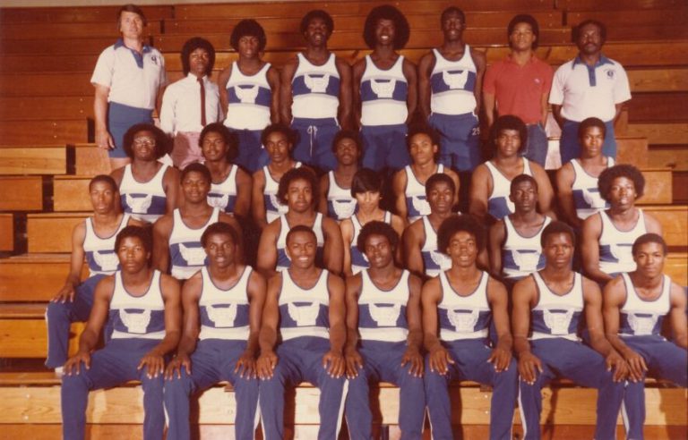 Track team in blue and white uniforms with coaches posed in rows on wooden bleachers, from Percy Hines' collection